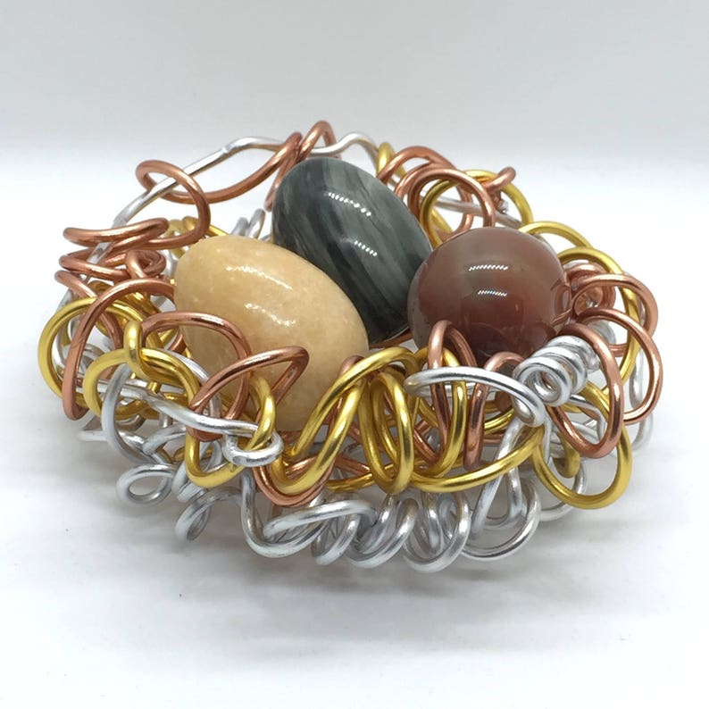 May include: A wire nest made of silver, copper, and gold wire holds three gemstone eggs. The eggs are a light brown, a dark green with white streaks, and a reddish brown.