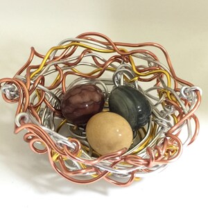 May include: A wire nest made of silver, copper, and gold wire with three colorful stones inside. The stones are brown, green, and beige.
