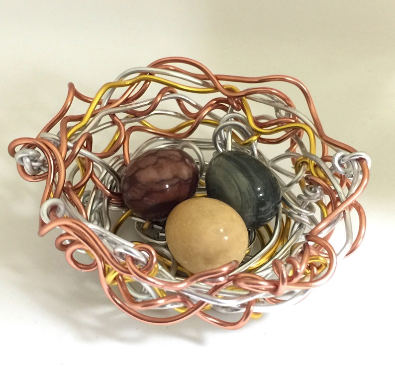 May include: A wire nest made of silver, copper, and gold wire with three colorful stones inside. The stones are brown, green, and beige.