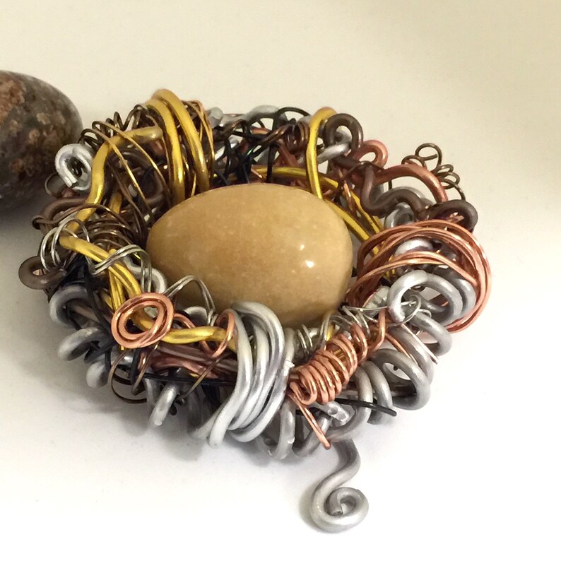 May include: A wire nest made of gold, silver, and copper wire with a tan stone egg in the center.
