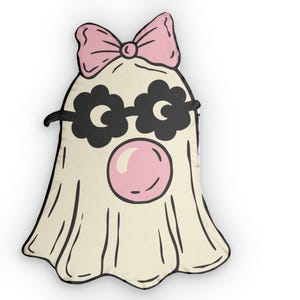 Peut inclure: Un coussin en forme de fantôme de couleur crème avec un nœud rose sur le dessus, des lunettes noires en forme de fleur et une bulle de chewing-gum rose. Le motif est souligné en noir, créant une illustration de style dessin animé.
