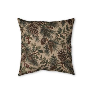 May include: A square throw pillow featuring a beige fabric base with a printed pattern of dark green pine needles, brown pinecones, and clusters of small red berries. The design covers the entire surface of the pillow in a repeating botanical motif.