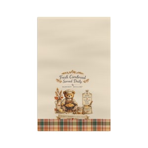 May include: A cream-colored waffle-weave kitchen towel featuring a central illustration of a teddy bear wearing an apron, holding a bowl of cornbread. Text reads Fresh Cornbread Served Daily by Harvest Hollow and Made With Love From Our Home Yours.