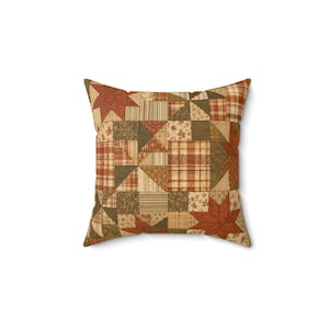May include: A square throw pillow featuring a patchwork design with various fabric patterns in shades of rust orange, olive green, and cream. The quilt-style cover displays geometric shapes, plaid, stripes, and floral motifs arranged in a grid.