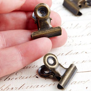 May include: A collection of small, antique brass-toned binder clips. One is held in a hand, with others on a background of handwritten script. The clips feature a classic design with a spring-loaded mechanism.