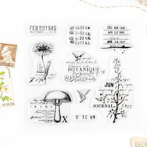 May include: A set of eight clear rubber stamps with black ink. The stamps feature various designs, including a mushroom, a flower, a bird, and a botanical illustration. The stamps also include text, such as "FEB 22 1945", "OCT 2653 GW", "DEC 14 1936", "JUN", and "JOURNAL".