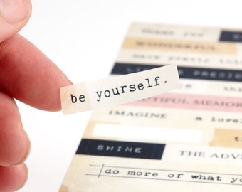 May include: A close-up of a white paper strip with the text "be yourself." printed in black typewriter font. The strip is held by a finger against a background of other paper strips with various text and designs.