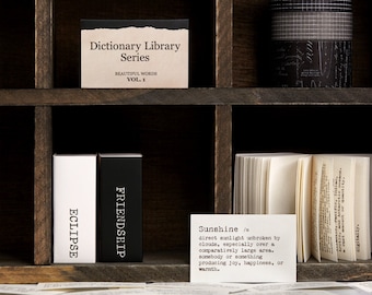 Könnte beinhalten: Ein Holzregal mit drei Reihen von Fächern. In der oberen Reihe befindet sich ein kleines Buch mit dem Titel "Dictionary Library Series" und den Worten "Beautiful Words Vol. 1". In der mittleren Reihe befindet sich ein Stapel schwarz-weiß gemusterten Washi-Bands. In der unteren Reihe befinden sich zwei kleine Bücher mit den Titeln "Eclipse" und "Friendship". Eine kleine Karte mit dem Wort "Sunshine" und einer Definition darunter ist ebenfalls vorhanden.