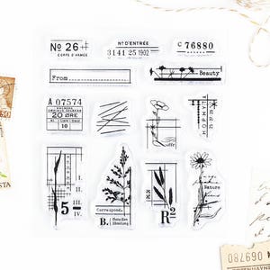 May include: A set of 10 clear rubber stamps with black ink featuring various designs, including botanical illustrations, text, and numbers. The stamps are arranged in a grid pattern on a white background.