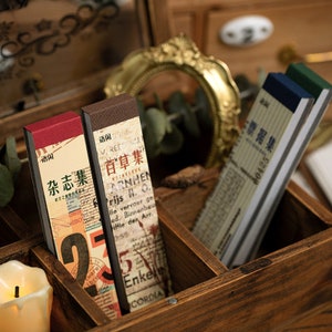 Peut inclure: Trois journaux en papier de style vintage avec des couvertures brunes, rouges et bleues. Les couvertures présentent du texte dans une langue étrangère et un collage d'images. Les journaux sont disposés dans une boîte en bois.