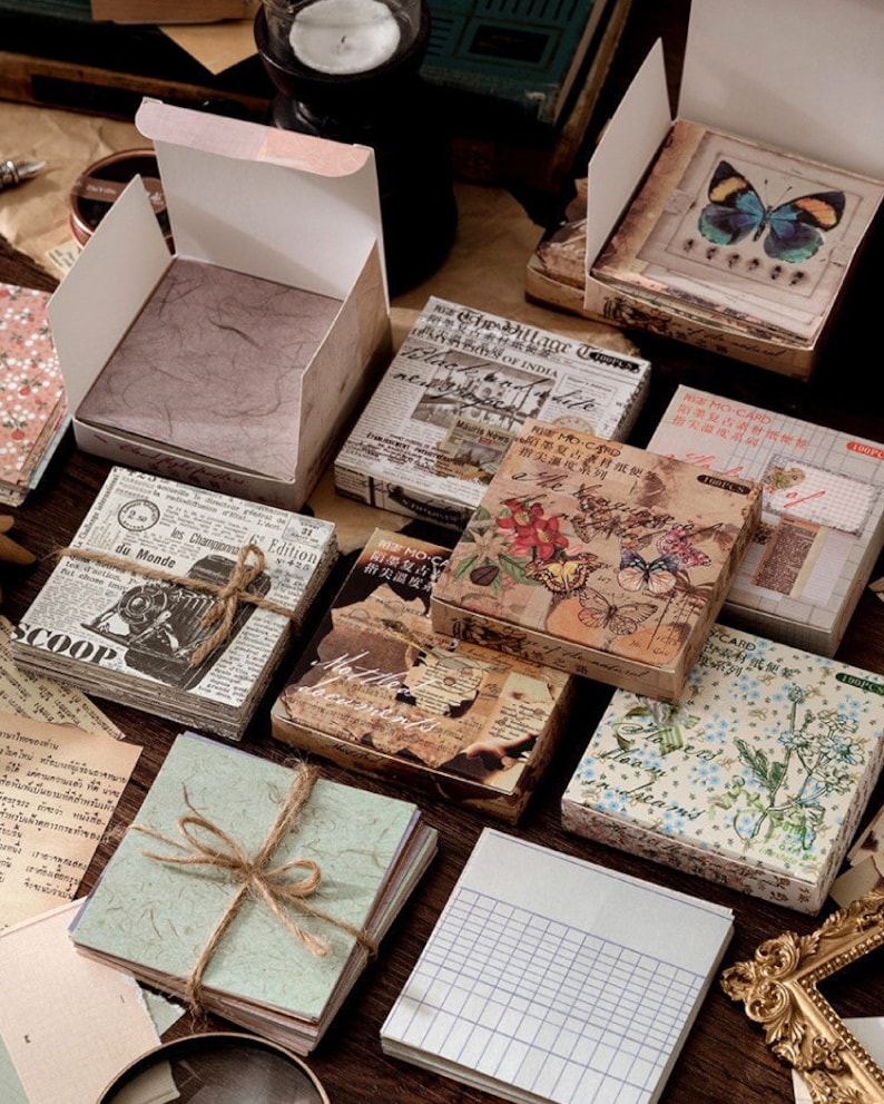 May include: Assortment of vintage-style decorative paper pads with floral, butterfly, and newspaper designs. The pads are packaged in boxes with a rustic aesthetic. The boxes have text in Japanese and English.