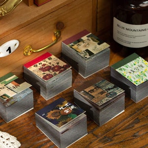 May include: Six small, square, stackable notebooks with different floral and patterned covers. The notebooks are arranged on a wooden surface. The covers are in various colors, including green, red, and blue.