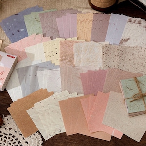 May include: A set of 30 decorative paper sheets with a variety of textures and colors, including beige, brown, pink, and green. The paper sheets are arranged in a grid pattern and have a rustic, handmade look. The text on the packaging reads "Hand Made Feathers".