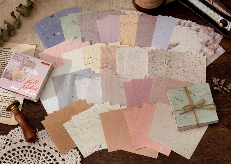 May include: A set of 30 decorative paper sheets with a variety of textures and colors, including beige, brown, pink, and green. The paper sheets are arranged in a grid pattern and have a rustic, handmade look. The text on the packaging reads "Hand Made Feathers".