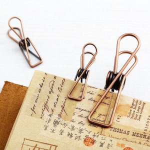 May include: Three antique copper binder clips of different sizes on a piece of paper with handwritten text and a brown leather background.