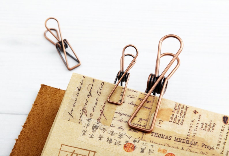 May include: Three antique copper binder clips of different sizes on a piece of paper with handwritten text and a brown leather background.
