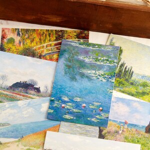 May include: A selection of art prints depicting impressionist paintings. The prints display scenes of water lilies, landscapes, and cityscapes, with a focus on vibrant colours and brushstrokes. The prints are displayed in a wooden box.