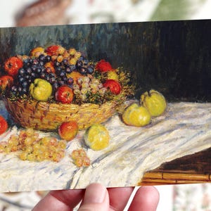 May include: A postcard depicting a still life painting of a basket of fruit. The basket contains red apples, purple and green grapes, and yellow quinces. The painting is on a white tablecloth.