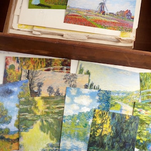 May include: A selection of art postcards depicting landscape paintings, including gardens, water lilies, and a windmill in a field of flowers. The postcards are stacked in a wooden drawer with aged paper.
