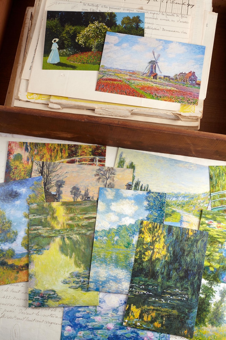 May include: A selection of art postcards depicting landscape paintings, including gardens, water lilies, and a windmill in a field of flowers. The postcards are stacked in a wooden drawer with aged paper.