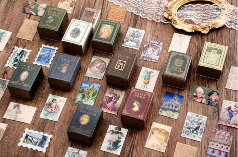 May include: A collection of vintage-style decorative boxes with various designs and titles, including "Notes on Nature", "Childood", "The Fred Factor", "Flower Fairy", and "Victoria". The boxes are made of cardboard and have a distressed, antique look. They are arranged on a wooden surface with other decorative elements, such as vintage postcards and stamps.