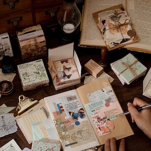 May include: A collection of decorative paper goods with floral and butterfly designs. The items include a set of decorative paper, a set of stickers, a set of washi tape, and a set of decorative paper clips. The items are arranged on a wooden table with a notebook and a pen.