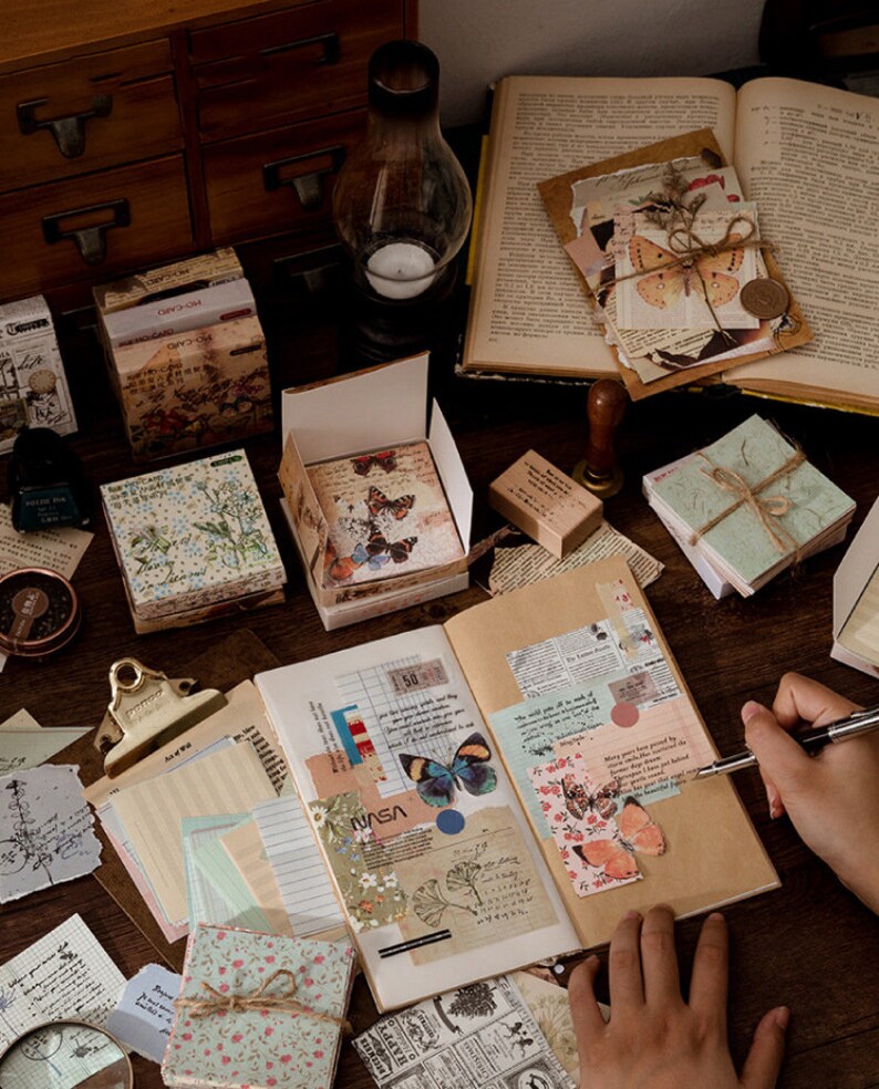 May include: A collection of decorative paper goods with floral and butterfly designs. The items include a set of decorative paper, a set of stickers, a set of washi tape, and a set of decorative paper clips. The items are arranged on a wooden table with a notebook and a pen.