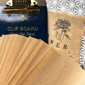 May include: A stack of brown parchment paper sheets with a gold clip board and a printed sheet of paper with a flower illustration and the word "FLOWER" on a wooden surface.
