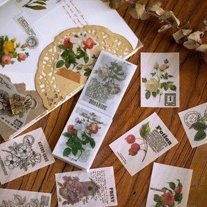 May include: A collection of vintage-style paper ephemera featuring floral and botanical illustrations. Includes stickers, labels, and decorative paper with roses, botanical prints, and text elements. The items are arranged on a wooden surface.