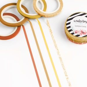 May include: A collection of washi tape rolls in warm tones, including rust, gold, and tan. The image features several rolls and strips of tape, with a stack of rolls labeled "wintertime" with floral accents.
