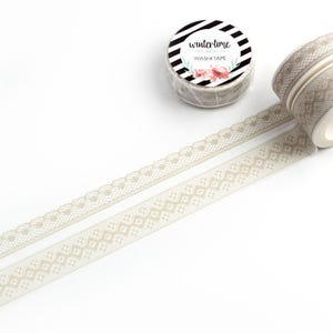 May include: Two rolls of beige washi tape with delicate lace patterns. The tape is unrolled to show the design. The roll of tape has a black and white label that says "wintertime washi tape".