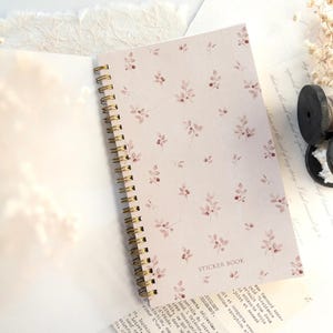 May include: A spiral-bound sticker book with a floral pattern in pink and burgundy. The open book reveals blank pages, while the cover displays the words "STICKER BOOK". The book is on a white surface with decorative elements.