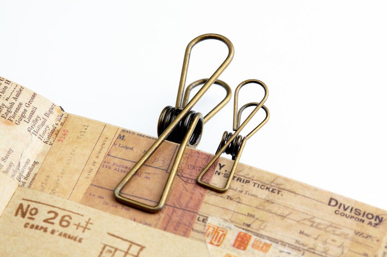 May include: Two antique gold binder clips, one larger than the other, are holding down a stack of vintage paper with printed text. The text includes the words "No 26++" and "CORPS D'ARM&Eacute;E".