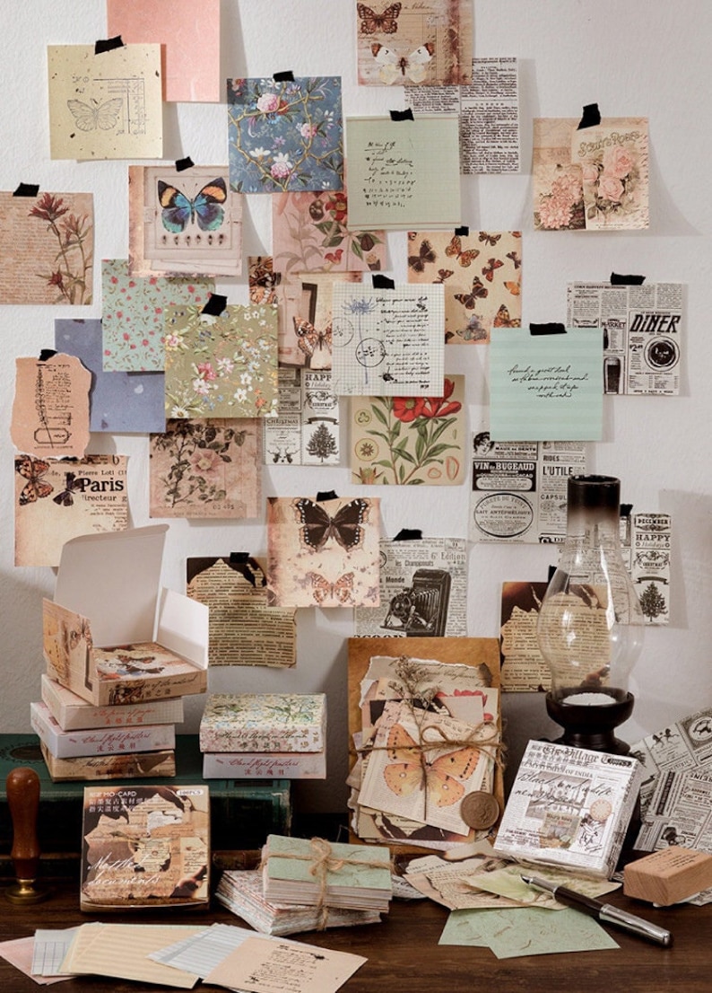 May include: A collection of vintage-style paper ephemera, including decorative papers, cards, and stickers, featuring butterflies, floral patterns, and text. The papers are arranged on a white wall and a wooden table, with some of the papers in boxes. The papers are perfect for scrapbooking, journaling, and other craft projects.