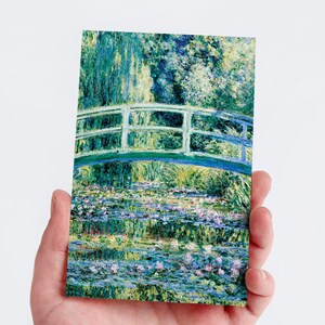May include: A postcard depicting a painting of a Japanese bridge over a pond with water lilies. The artwork uses green, blue, and purple hues. The bridge has a white railing and arches over the water, reflecting in the pond.