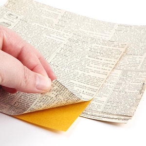 May include: A hand holding a stack of old, yellowed newspaper pages. The top page is being peeled back to reveal a yellow piece of paper underneath.
