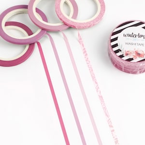 May include: A collection of pink washi tape rolls and strips on a white surface. The rolls vary in shades of pink, with one roll featuring the text "wintertime" and floral accents. The strips of tape are in varying shades of pink.