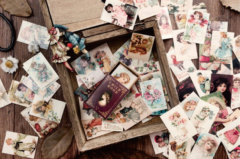 May include: A wooden box filled with vintage-style postcards featuring children. The box is open and the postcards are arranged in a random pattern. The box is labeled "CHILDHOOD".