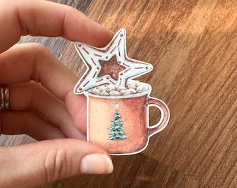 May include: A watercolor illustration of a mug of hot chocolate topped with marshmallows and a star-shaped cookie. The mug has a Christmas tree design. The cookie is light blue with red accents and a brown star center.