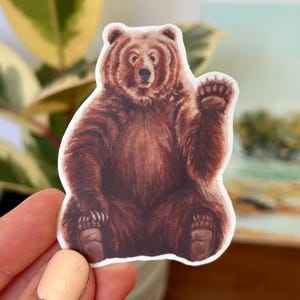 May include: A brown bear sticker with a friendly expression, waving with its right paw. The bear is sitting on its haunches with its paws visible. The sticker is on a white background.