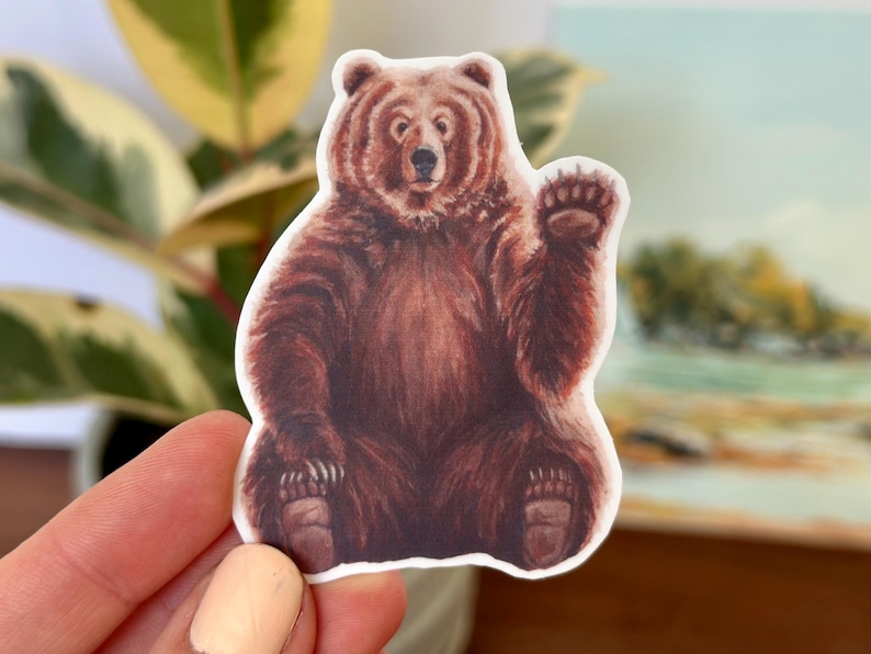 May include: A brown bear sticker with a friendly expression, waving with its right paw. The bear is sitting on its haunches with its paws visible. The sticker is on a white background.