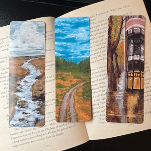 Könnte beinhalten: Drei Aquarell-Lesezeichen mit verschiedenen Landschaften. Das erste Lesezeichen zeigt einen blauen Bach, der durch ein grünes Tal fließt. Das zweite Lesezeichen zeigt eine unbefestigte Straße, die sich durch einen Wald schlängelt. Das dritte Lesezeichen zeigt eine Straßenszene mit einer alten Straßenbahn und einem Gebäude im Hintergrund.