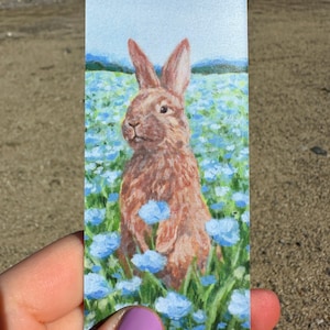May include: A brown rabbit with white fur on its chest stands in a field of blue flowers. The rabbit is looking to the left of the image. The background is a blue sky with white clouds.