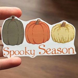 Op de afbeelding: Een sticker met drie pompoenen in groen, oranje en geel. De tekst "Spooky Season" staat in oranje letters eronder. De sticker wordt door een hand vastgehouden.