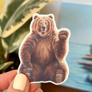 May include: A brown bear sticker with a friendly expression, waving with its right paw. The bear is sitting on a white background.