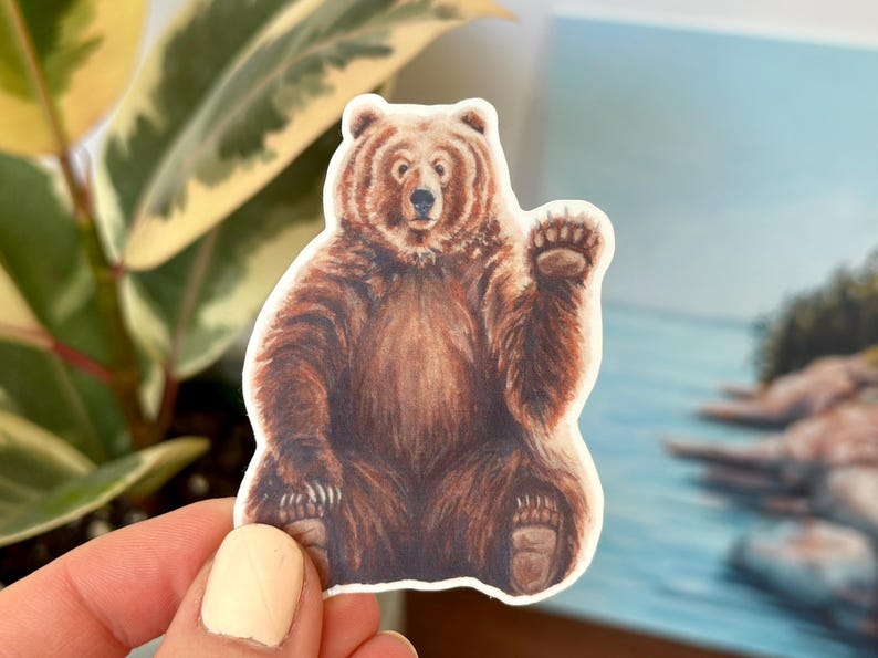 May include: A brown bear sticker with a friendly expression, waving with its right paw. The bear is sitting on a white background.