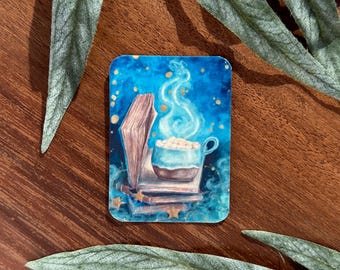May include: A blue and gold sticker with a watercolor illustration of a mug of hot chocolate with marshmallows, steam rising from the mug, and a stack of books. The mug is sitting on top of the books.