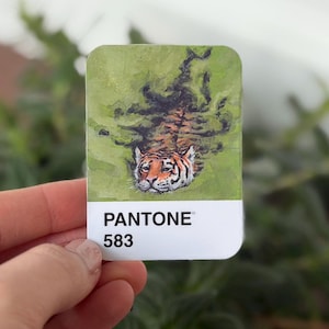 May include: A rectangular sticker with a tiger illustration on a green backdrop. The tiger's head and shoulders are visible, with orange and black stripes. The sticker has a white border with the word "PANTONE" and the number "583".