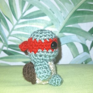 May include: A small crocheted turtle toy with a red bandana and black eyes. The turtle is sitting on a green surface.