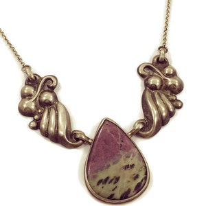 Op de afbeelding: Een zilveren ketting met een traanvormige hanger van groene en roze steen. De hanger is omgeven door twee zilveren swirls.